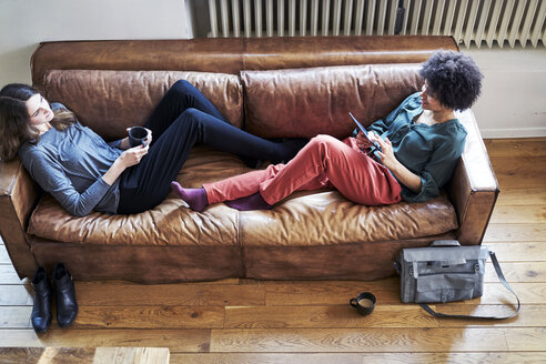 Zwei junge Frauen liegen auf einer Couch mit einer Tasse Kaffee und einem Tablet - FMKF04802