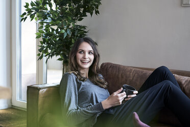 Happy young woman lying on couch with cup of coffee - FMKF04803