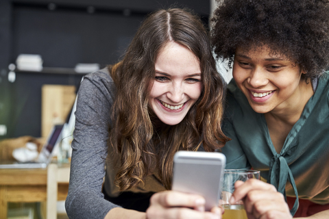 Zwei lächelnde junge Frauen teilen sich ein Mobiltelefon im Büro, lizenzfreies Stockfoto