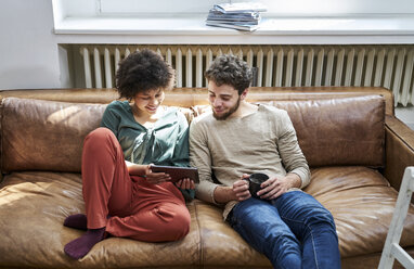 Young man and young woman sitting on couch with cup of coffee and tablet - FMKF04831