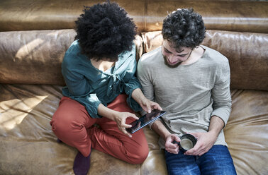 Young man and young woman sitting on couch with cup of coffee and tablet - FMKF04832