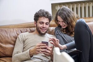 Junger Mann und junge Frau sitzen auf der Couch und teilen sich Handy und Kopfhörer - FMKF04852