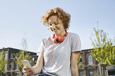 Lächelnde junge Frau mit Kopfhörern und Smartphone in städtischer Umgebung - PDF01417