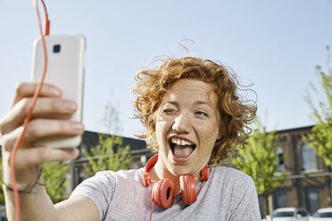 Verspielte junge Frau mit Kopfhörern macht ein Selfie in städtischer Umgebung - PDF01418