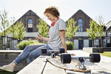 Lächelnde junge Frau mit Kopfhörern und Longboard, die ihr Smartphone in einer städtischen Umgebung benutzt - PDF01420