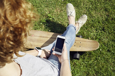 Junge Frau mit Smartwatch und Longboard nutzt Smartphone auf einer Wiese - PDF01425