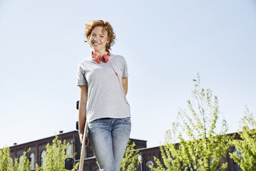 Lächelnde junge Frau mit Longboard und Kopfhörern in städtischer Umgebung stehend - PDF01427
