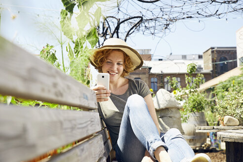 Smiling young woman wearing straw using smartphone in urban garden - PDF01439