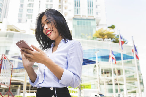 Thailand, Bangkok, lächelnde Geschäftsfrau in der Stadt, die auf ihr Handy schaut - WPEF00077