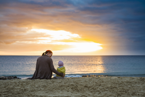 Mother with little daughter sitting on the beach at sunset stock photo