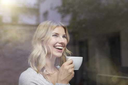 Porträt einer lachenden Frau mit einer Tasse Kaffee hinter einer Fensterscheibe - PNEF00519
