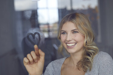 Porträt einer glücklichen blonden Frau, die ein Herz auf eine Fensterscheibe zeichnet - PNEF00528