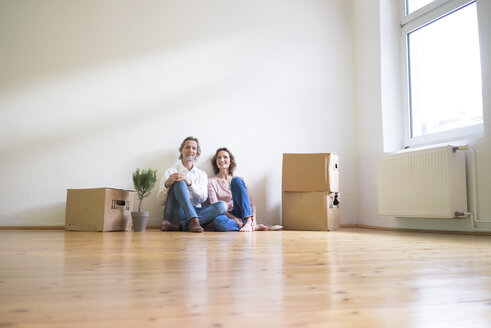 Smiling mature couple sitting on floor in empty room next to cardboard boxes - MOEF00741