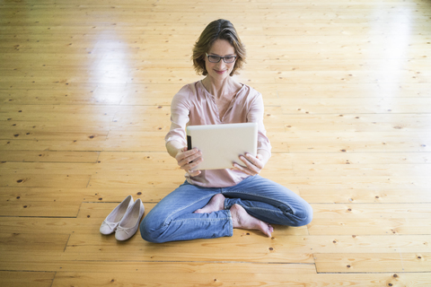 Smiling mature woman sitting on wooden floor taking selfie with tablet stock photo