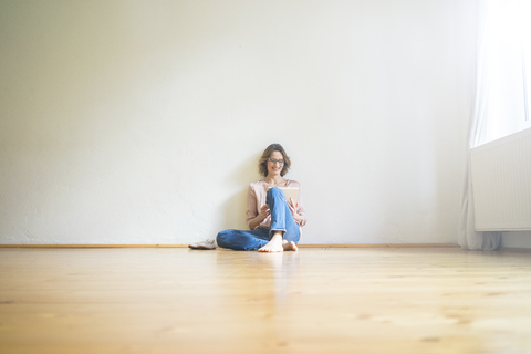 Lächelnde reife Frau, die in einem leeren Raum auf dem Boden sitzt und ein Tablet benutzt, lizenzfreies Stockfoto