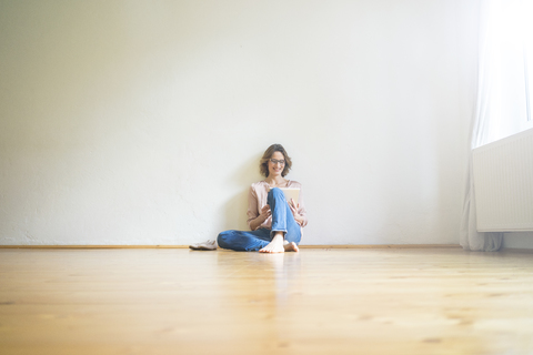 Smiling mature woman sitting on floor in empty room using tablet stock photo