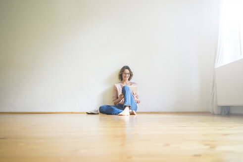 Lächelnde reife Frau, die in einem leeren Raum auf dem Boden sitzt und ein Tablet benutzt - MOEF00758