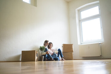 Mature couple sitting on floor in empty room next to cardboard boxes using tablet - MOEF00761