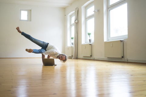 Mature man doing a handstand on floor in empty room looking at tablet stock photo