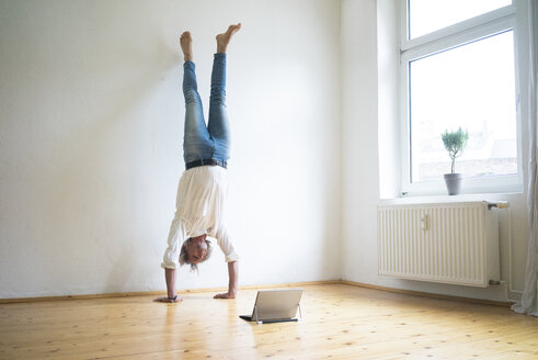 Älterer Mann macht einen Handstand auf dem Boden in einem leeren Raum und schaut auf ein Tablet - MOEF00767