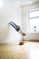 Älterer Mann macht einen Handstand auf dem Boden in einem leeren Raum und schaut auf ein Tablet - MOEF00768