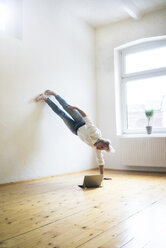Mature man doing a handstand on floor in empty room looking at tablet - MOEF00768