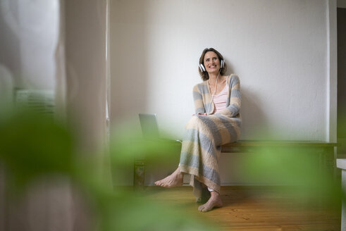 Smiling mature woman sitting on wooden bench at home listening to music - MOEF00785