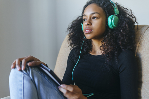 Porträt einer jungen Frau, die im Sessel sitzt und mit Kopfhörern und einem Tablet Musik hört, lizenzfreies Stockfoto