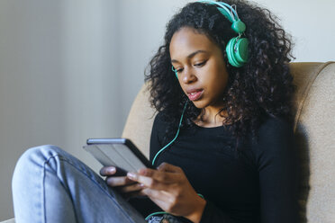 Porträt einer jungen Frau, die im Sessel sitzt und mit Kopfhörern und einem Tablet Musik hört - KIJF01906