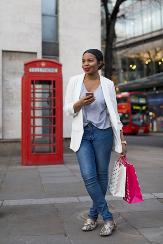 UK, London, lächelnde Frau mit Handy und Einkaufstüten in der Stadt, lizenzfreies Stockfoto