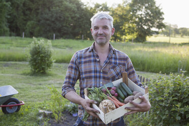 Porträt eines lächelnden reifen Mannes, der eine Kiste mit frisch geerntetem Gemüse im Garten trägt - FSIF00183