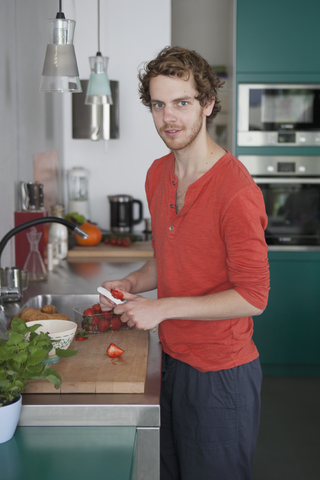 Porträt eines jungen Mannes beim Schneiden von Erdbeeren am Küchentisch, lizenzfreies Stockfoto