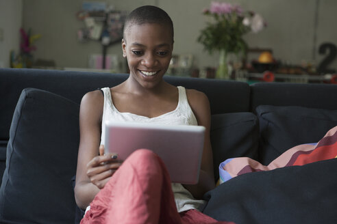 Lächelnde junge Frau, die auf dem Sofa zu Hause ein digitales Tablet benutzt - FSIF00226