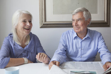 Happy senior couple with newspaper sitting at table in house - FSIF00264