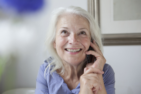 Happy senior woman looking up while answering mobile phone at home stock photo