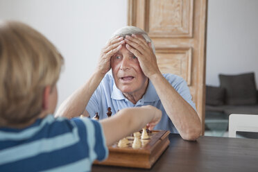 Shocked senior man with head in hands looking at grandson while playing chess at home - FSIF00274