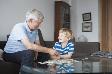 Happy grandson and grandfather looking at each other while solving jigsaw puzzle in living room - FSIF00284