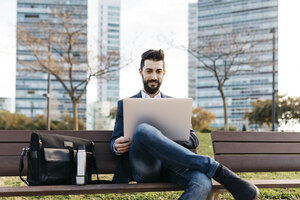 Geschäftsmann sitzt auf einer Bank vor einem Bürogebäude und benutzt einen Laptop - JRFF01540