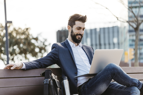 Businessman sitting on bench outside office building using laptop stock photo