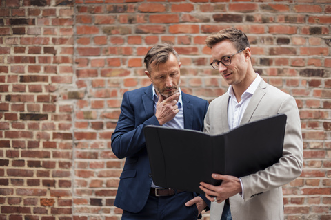 Two businessmen sharing folder at brick wall stock photo