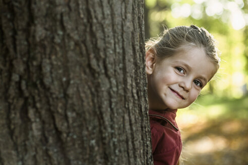 A young smiling girl peeking from behind a tree trunk - FSIF00522