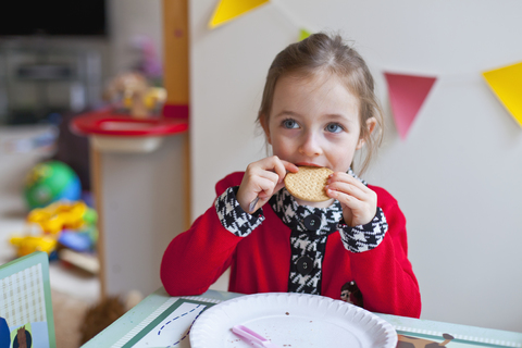 Ein kleines Mädchen sitzt an einem Tisch und beißt in einen Keks, lizenzfreies Stockfoto