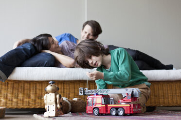 Junge spielt mit Spielzeug, Mutter und Vater liegen auf dem Sofa im Hintergrund - FSIF00790