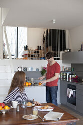 Frau sitzt am Esstisch, während ein Mann in der Küche das Frühstück vorbereitet - FSIF00800