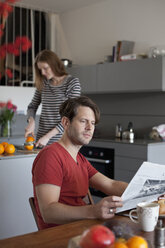 Mann liest in der Küche Zeitung, Frau macht im Hintergrund Orangensaft - FSIF00809