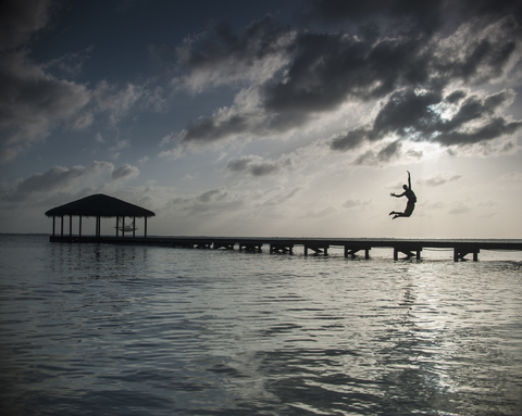 Silhouette person jumping into sea against cloudy sky at dusk stock photo