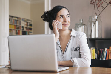 Beautiful young woman using mobile phone with laptop on table at home - FSIF00842