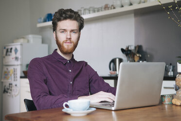 Porträt eines selbstbewussten jungen Mannes, der am Tisch in der Küche einen Laptop benutzt - FSIF00847