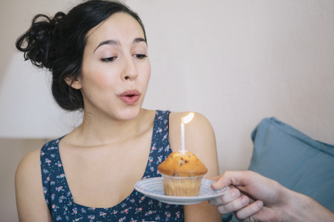 Junge Frau bläst die Kerze auf einem Kuchen aus, den ein Mann zu Hause hält, lizenzfreies Stockfoto
