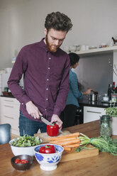 Young man chopping red bell pepper at kitchen table with woman in background - FSIF00863